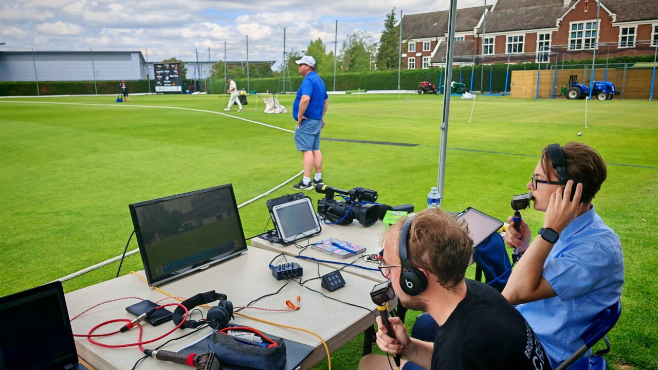 UoG students sat a table commentating on an England Seniors match with the green pitch in front of them and spectator in the background