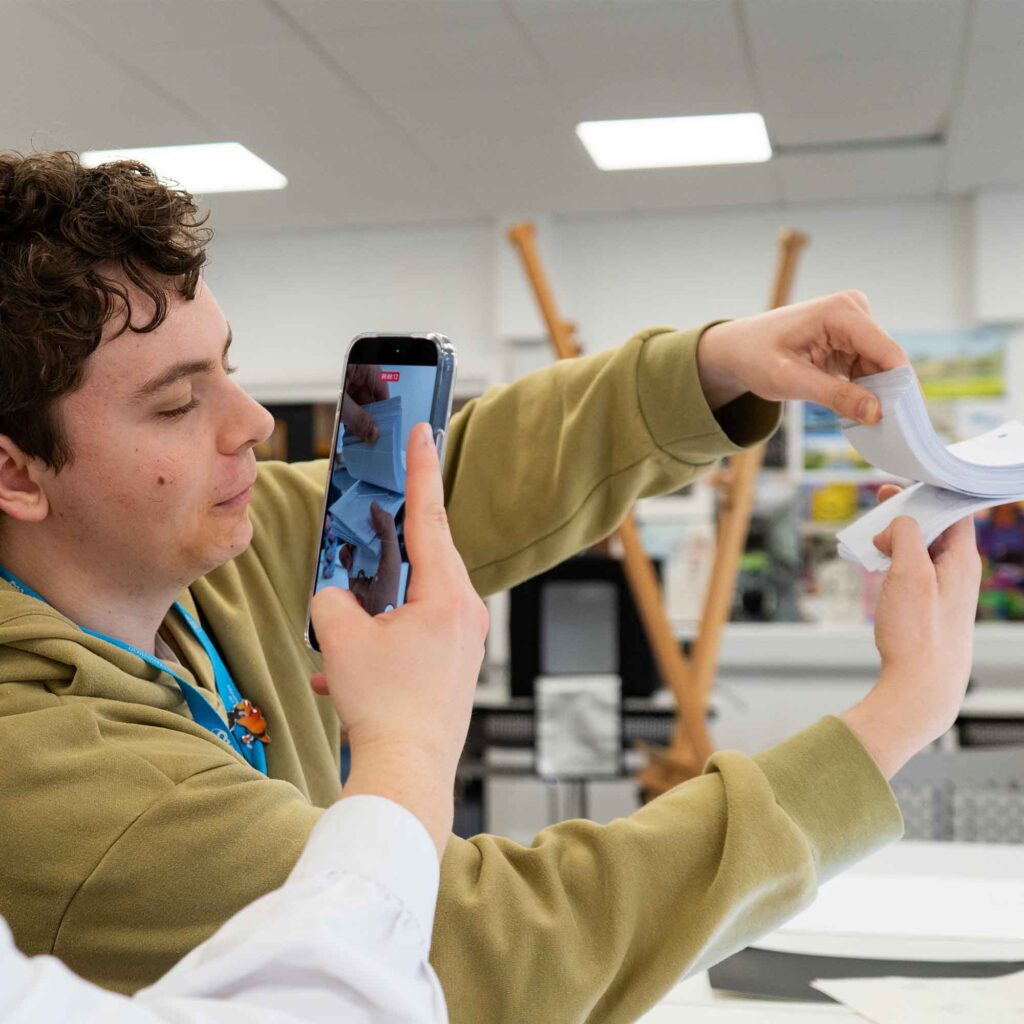 A student flipping a flipbook whilst someone else holds a phone recording it in front.