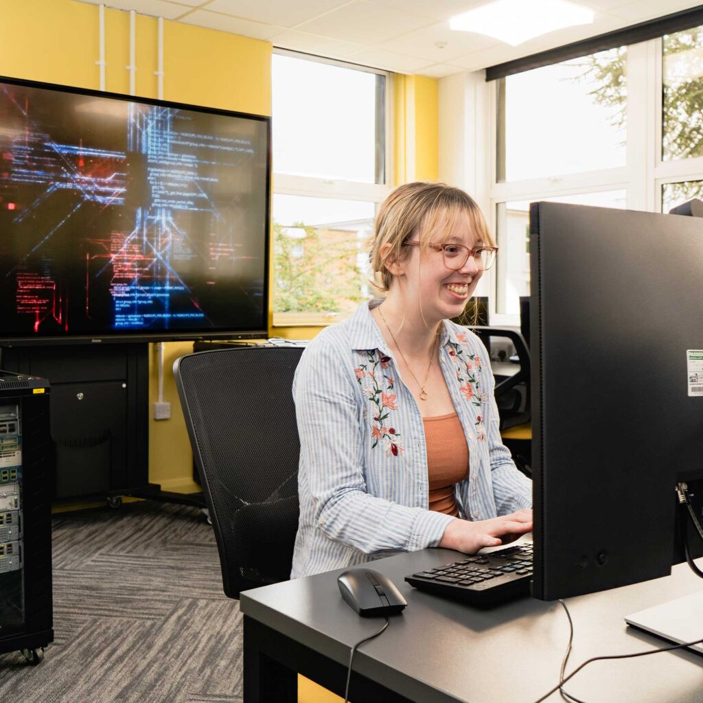 A student using computing equipment in a room with servers and a large digital screen.