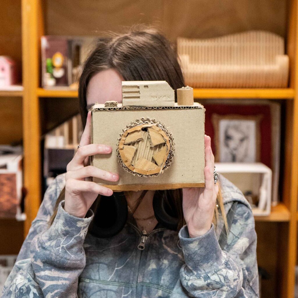 Student holding up a camera made of cardboard up in front of their face.
