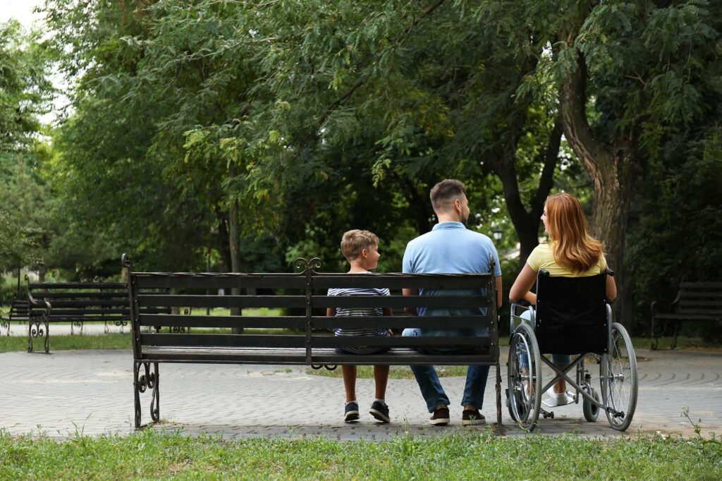 A woman in a wheelchair sits alongside a man and a child who sit on a bench in a park.