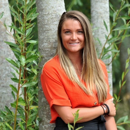 A photo of Alice smiling outside with greenery in the background.