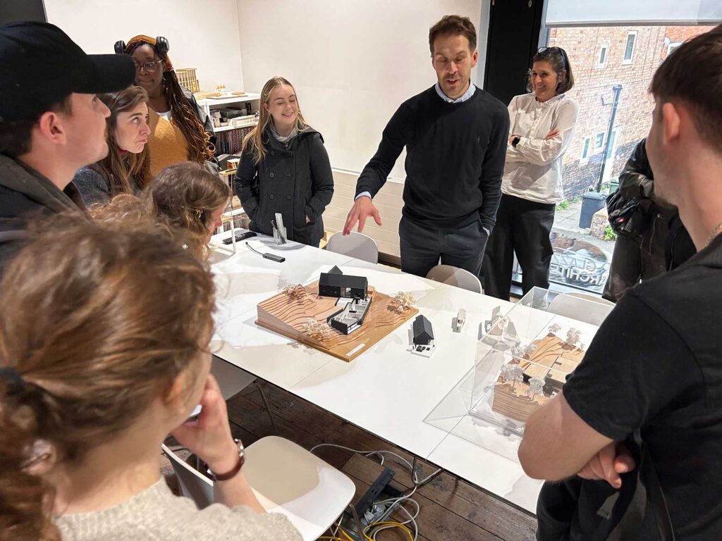 Students stand around a table with scale models being presented by a member of Glazzard Architects.