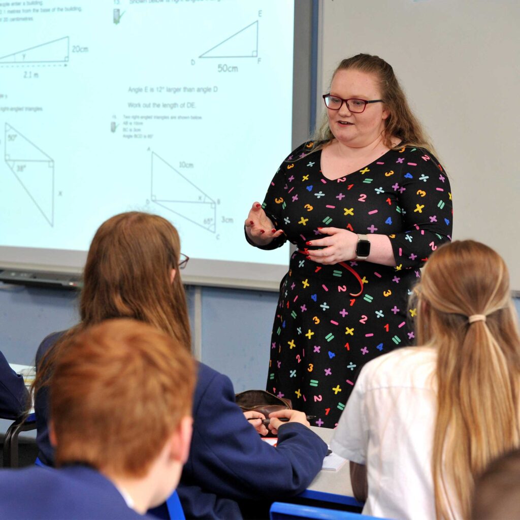 Teacher in a classroom with students facing the front of room which has a smart screen displaying triangles and angles.