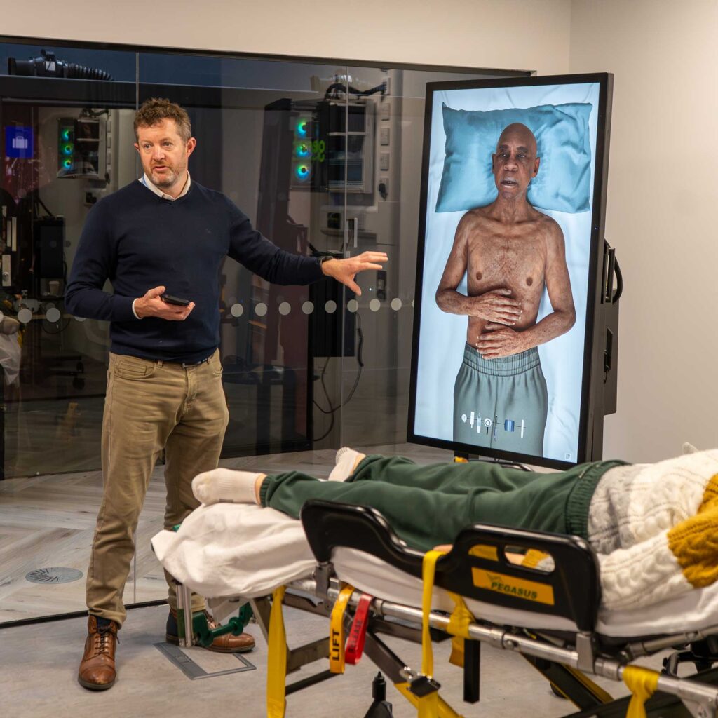A lecturer points towards a digital screen showing a patient lying on a bed in the immersive suite in FutureSim.