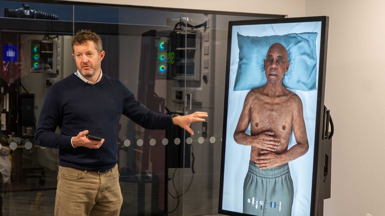 A lecturer points towards a digital screen showing a patient lying on a bed in the immersive suite in FutureSim.