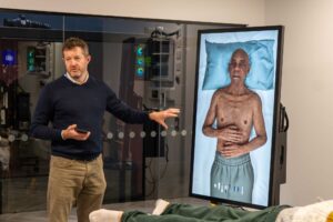 A lecturer points towards a digital screen showing a patient lying on a bed in the immersive suite in FutureSim.