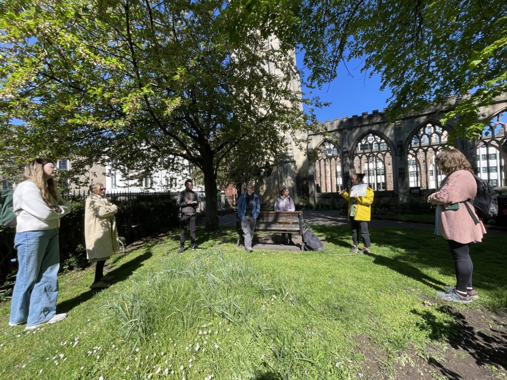 Seven people standing in a large circle within a church garden, with trees around them and a bench centre of the image.