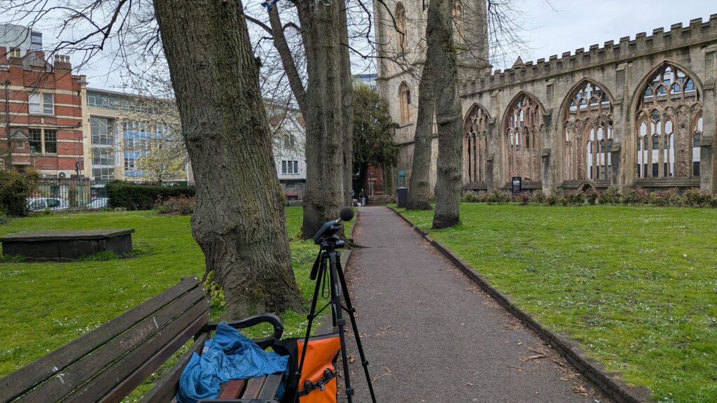 A concrete path leading through a green park space with a church on the right, other buildings on the left and a recording microphone on a tripod next to a bench in the foreground.