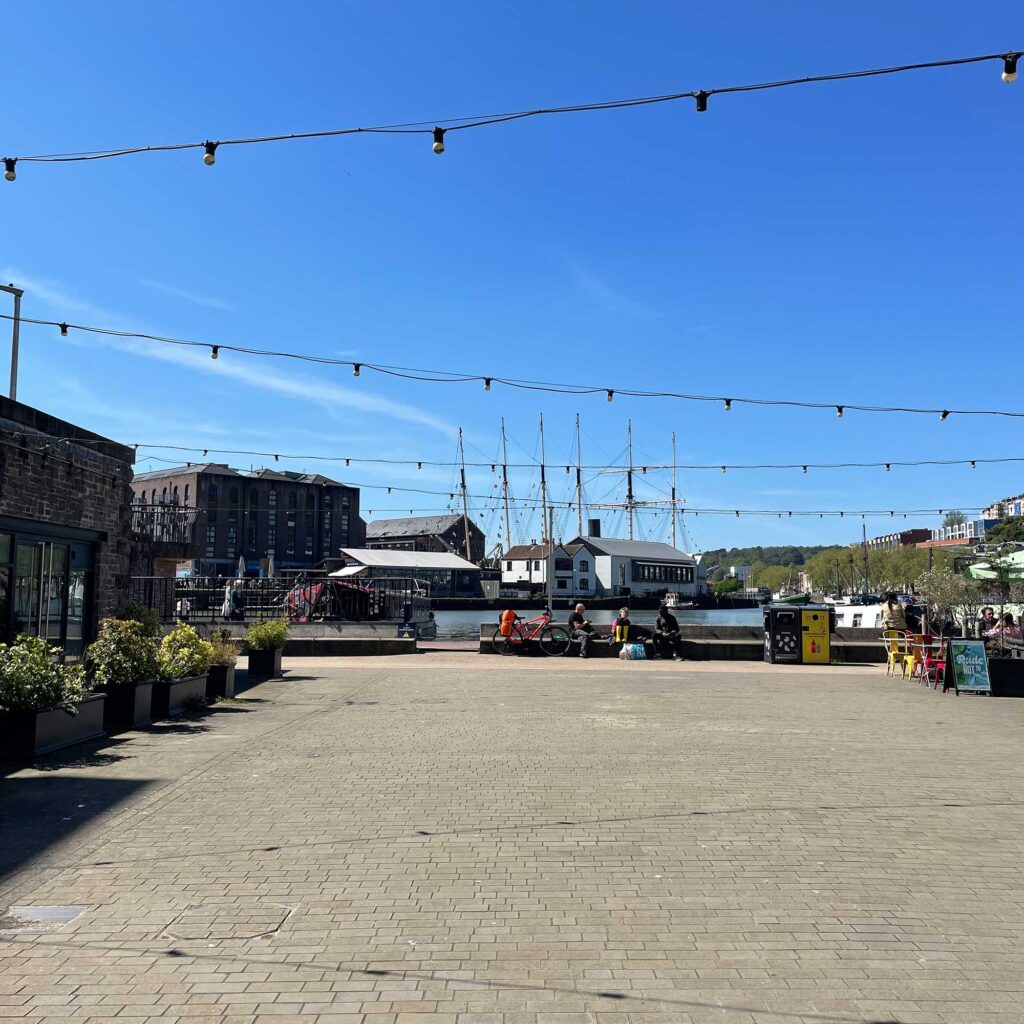 A paved promenade area next to a harbour with people sitting down and a tallship and buildings in the background.