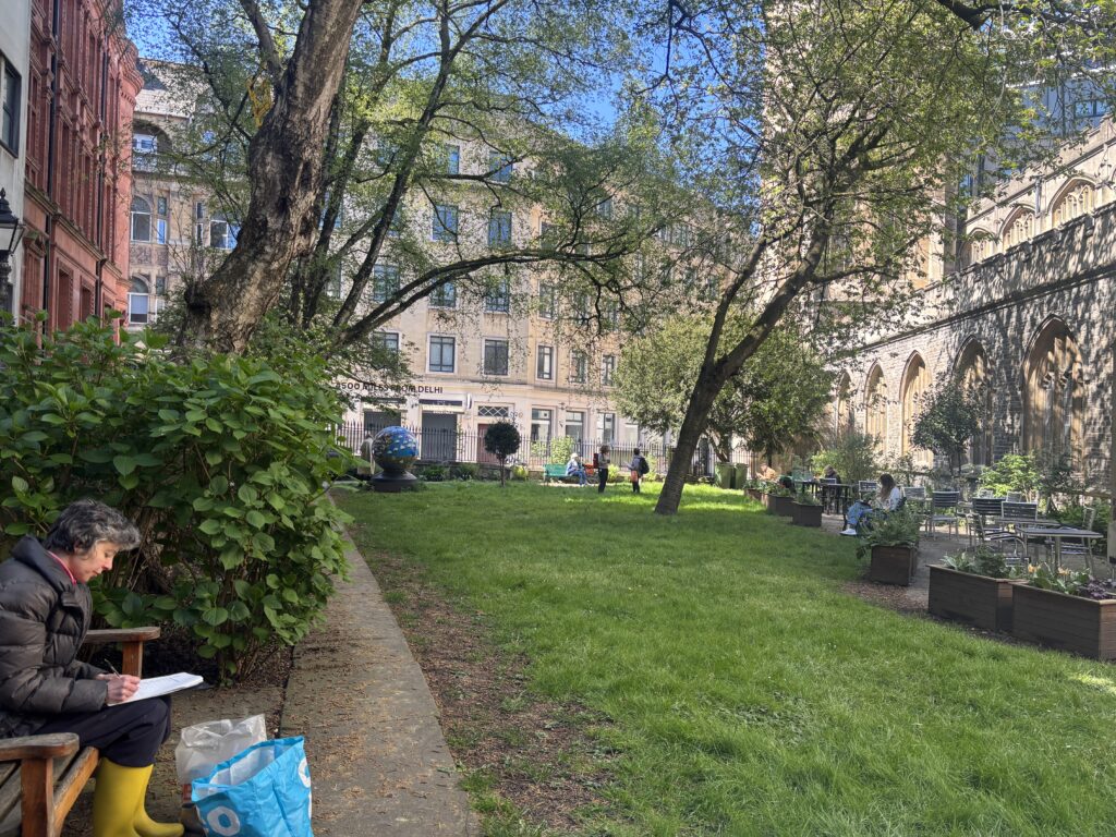 Green park space with someone writing on a bench in the foreground wearing yellow wellington boots and other people sat on tables and benches in the background.
