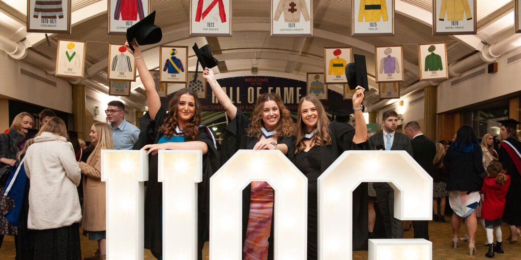 Three female students at Graduation in front of giant light up letters spelling UoG and holding their motar boards in the air.