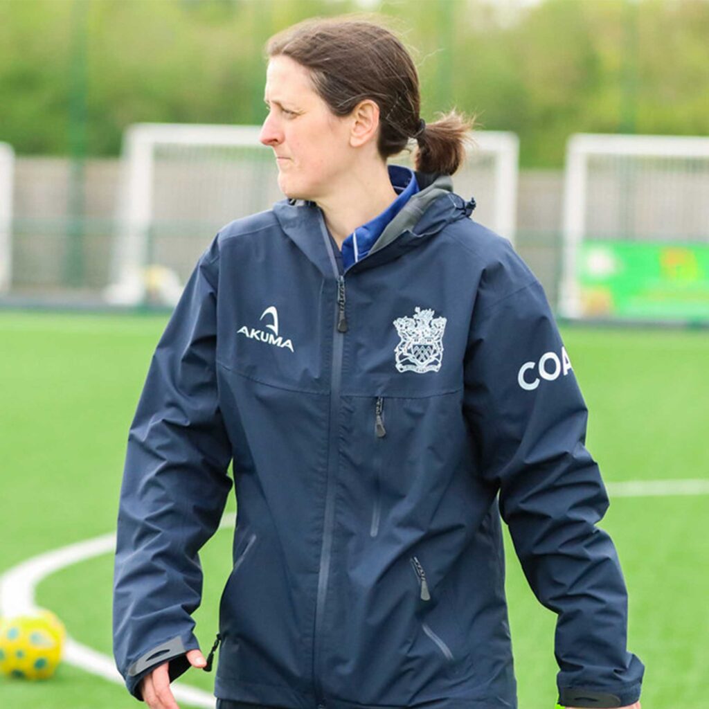 Kate Gumm wearing coach jacket walking on pitch with football goals in the background.