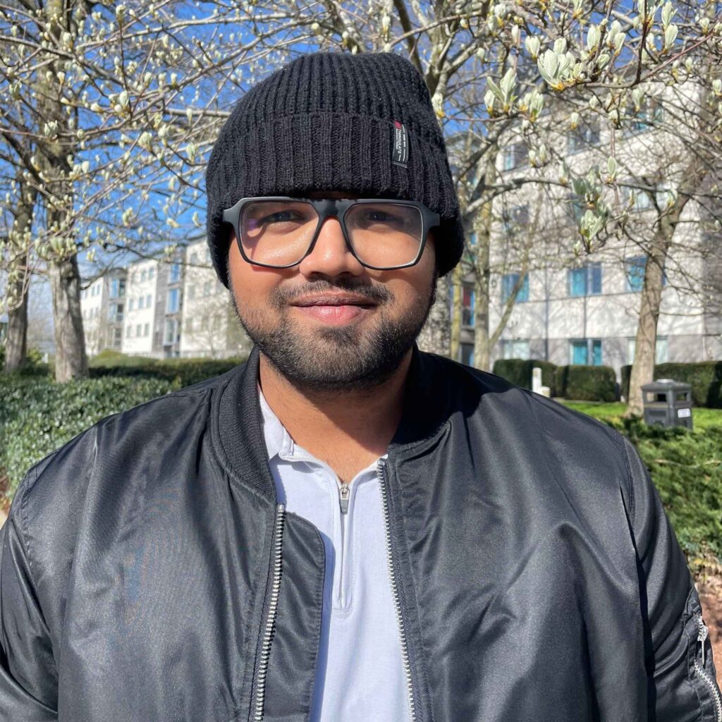 Head and shoulders photograph of Pavan wearing a beanie hat and glasses, with grass and trees with white blossom behind him