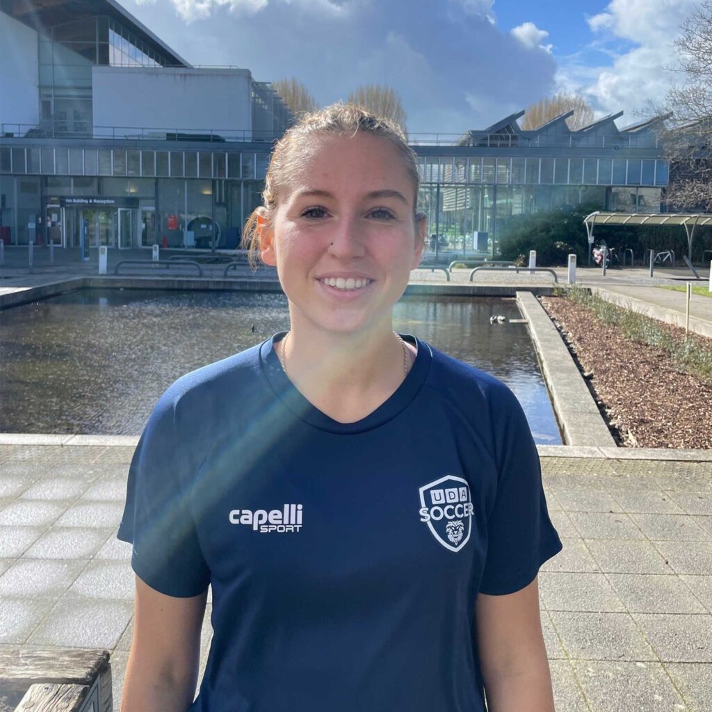 Head and shoulders photograph of Emma wearing a UDA soccer navy blue t-shirt, with a pond and Oxstalls main building in the background.