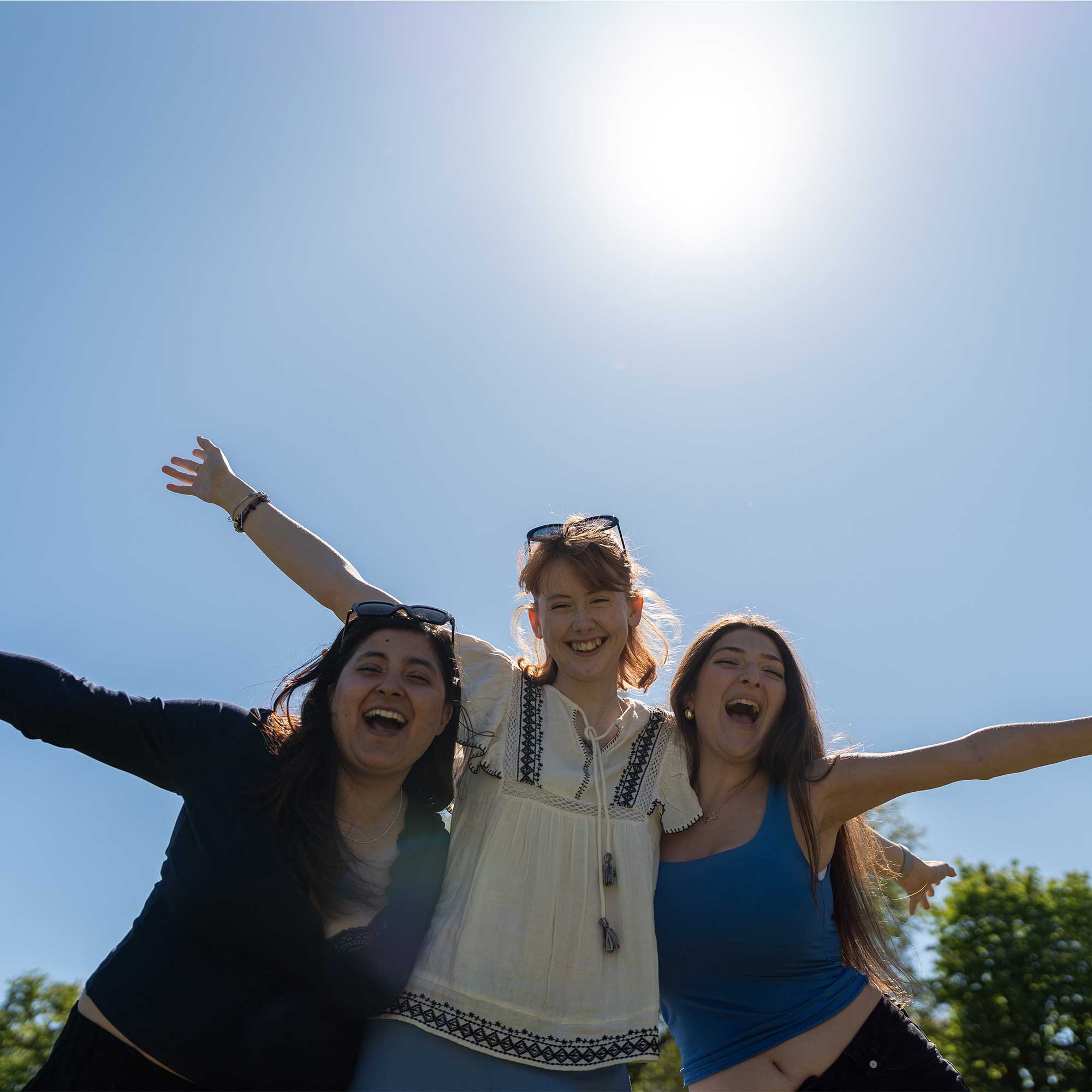 Three students standing close together, outdoors on a sunny day with a clear blue sky behind them and trees in the background. Each student has both arms lifted outward in a wide, open pose.