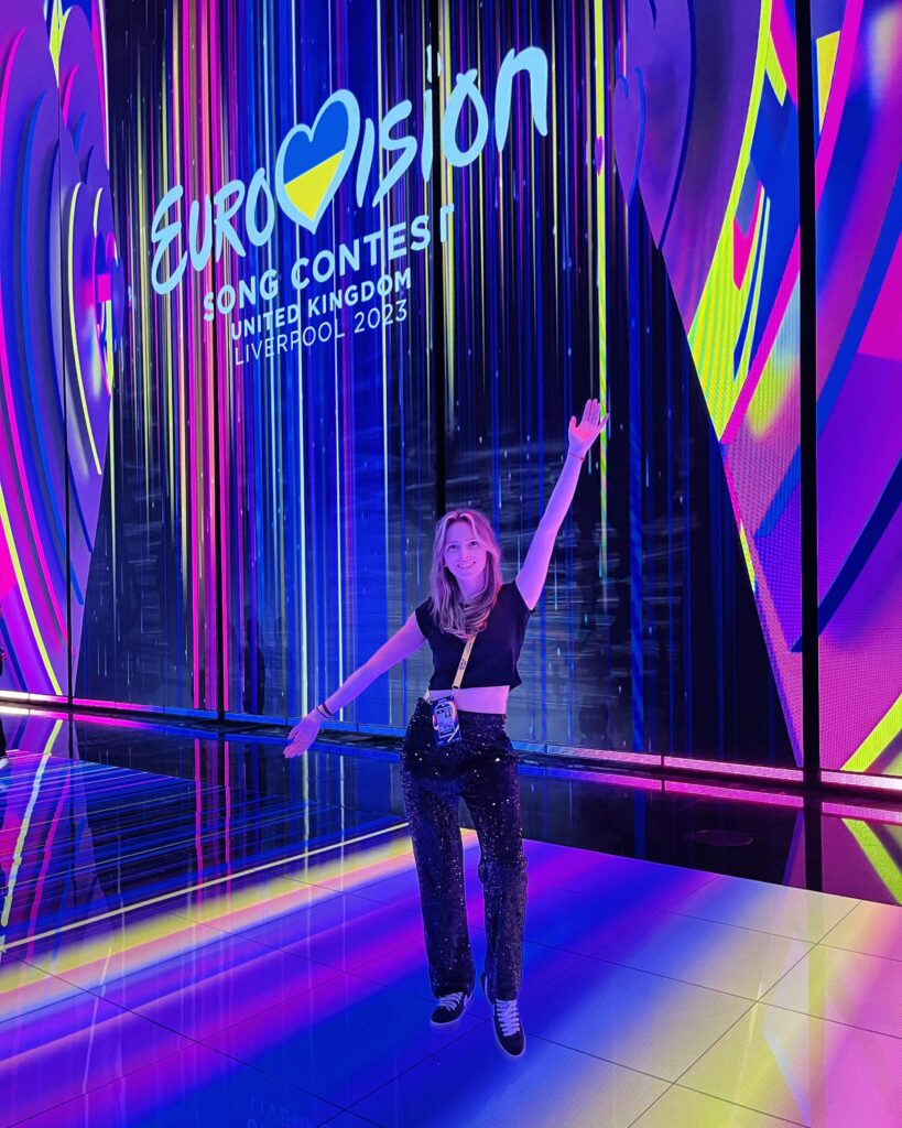 Anastazia posing in front of a colourful Eurovision Song Contest 2023 display in Liverpool, with bright neon lighting and reflective flooring.