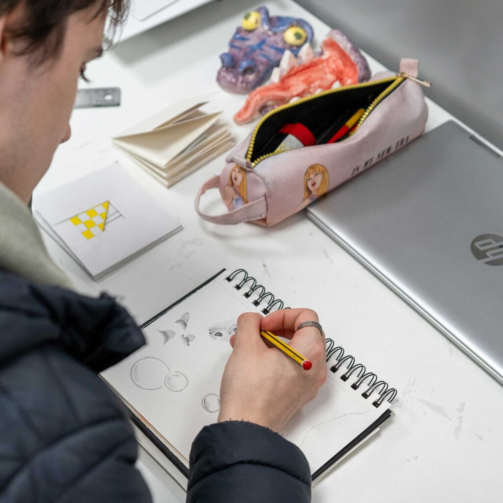 A student draws in a notebook surrounded by creative equipment.