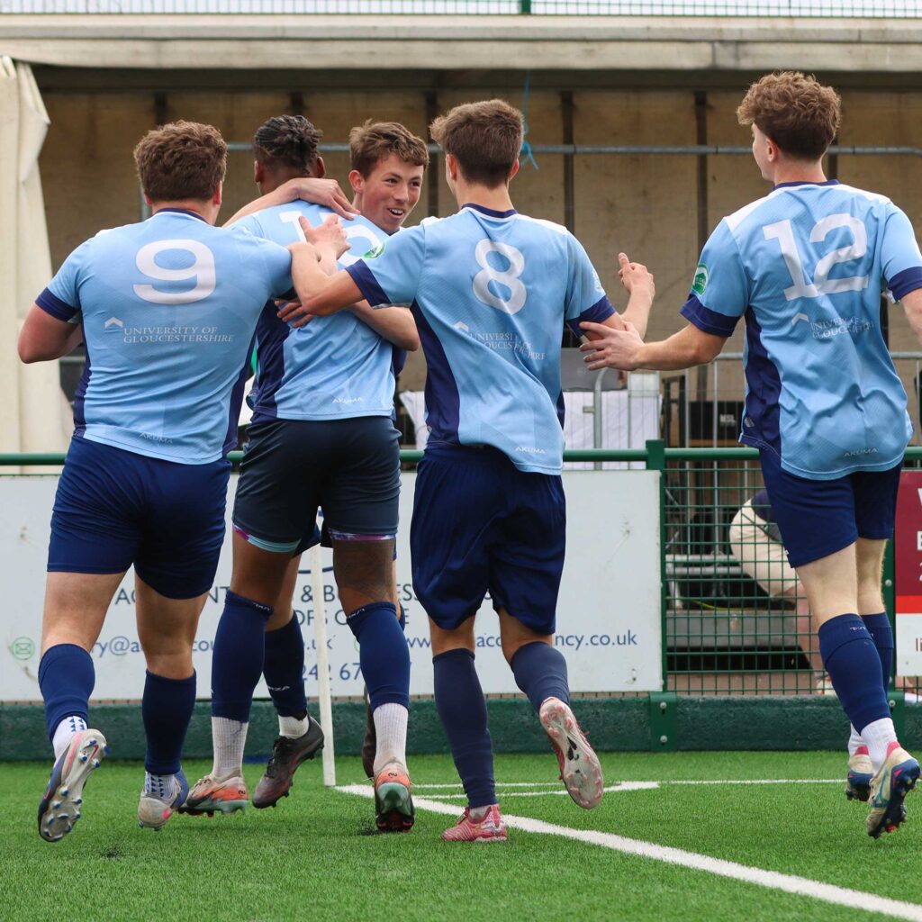 Mens football students running towards and hugging the goal scorer whilst celebrating a goal