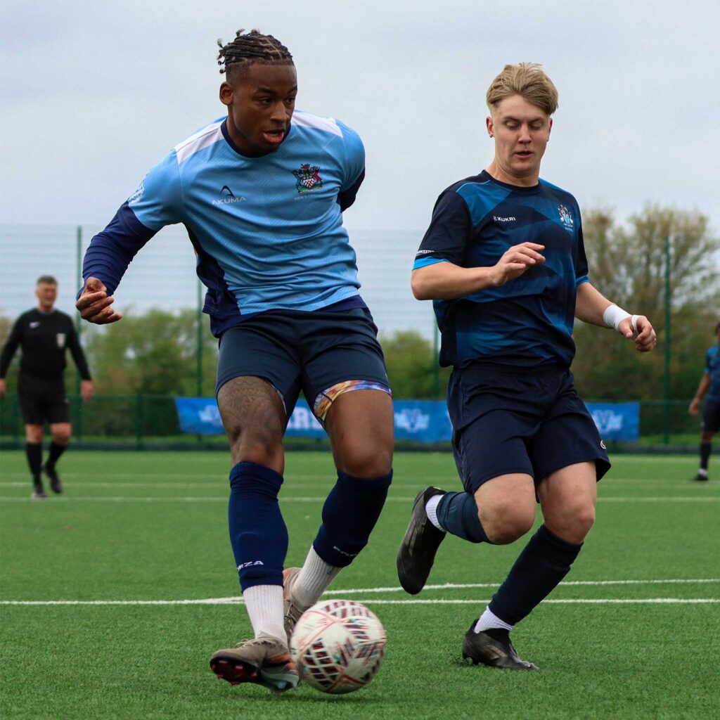 Two football players side by side on a pitch chasing the ball, with one player in a light blue kit and the other in a dark kit.
