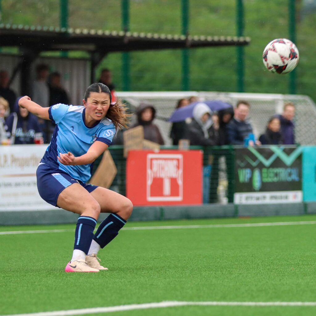 Female football player in a University of Gloucestershire football kit striking a ball during a match on a grass pitch with spectators blurred in the background.