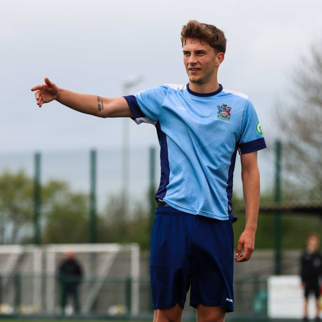 Male football player in a University of Gloucestershire football kit standing on a pitch, pointing with one arm, with blurred spectators in the background.