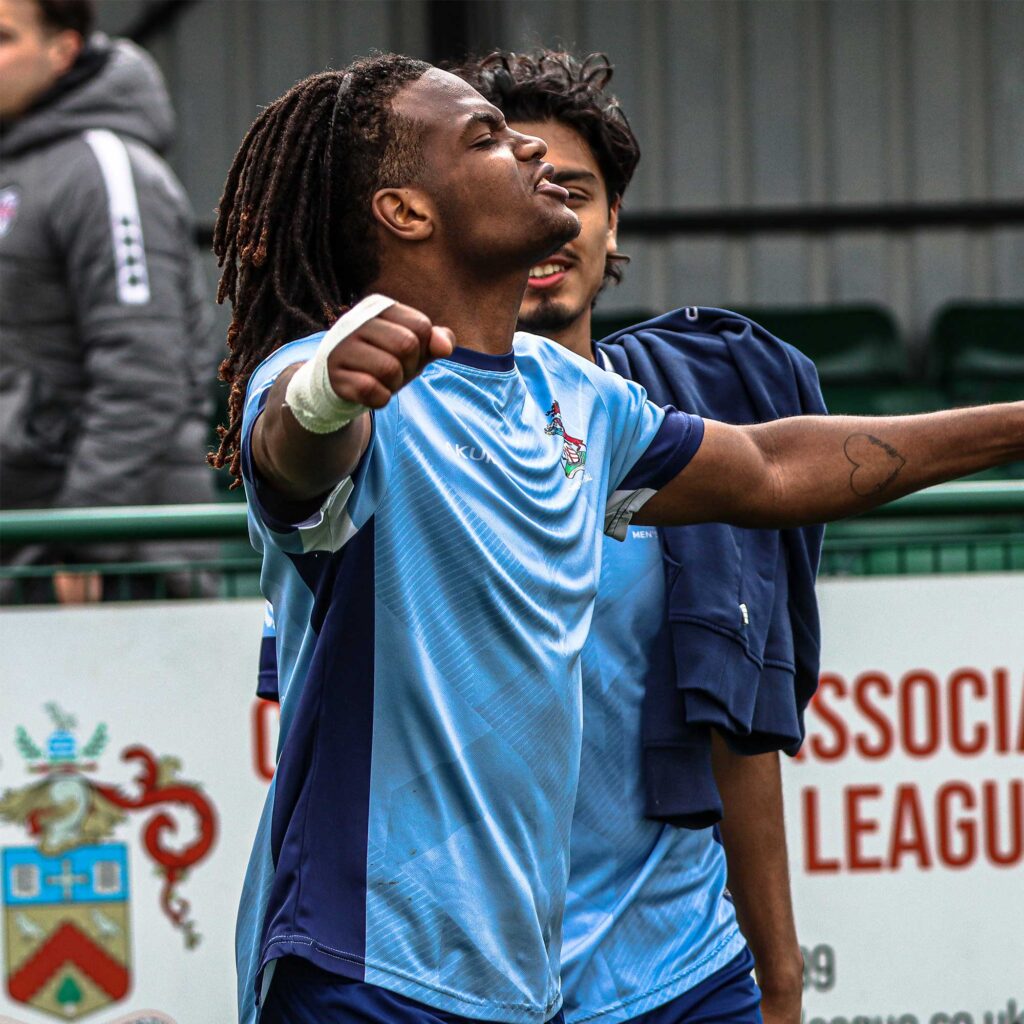 A male football player wearing a light blue University of Gloucester kit raises both arms in celebration on a pitch, with another teammate standing close behind.