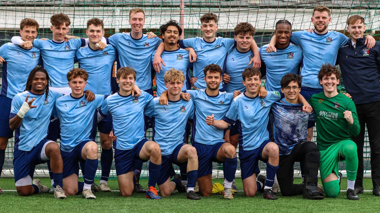 Eighteen male football players, wearing University of Gloucestershire kits, standing in two rows in front of a goal for a team photograph.