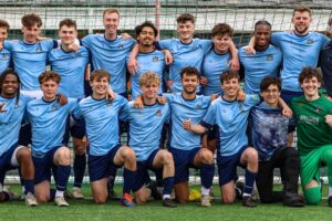 Eighteen male football players, wearing University of Gloucestershire kits, standing in two rows in front of a goal for a team photograph.