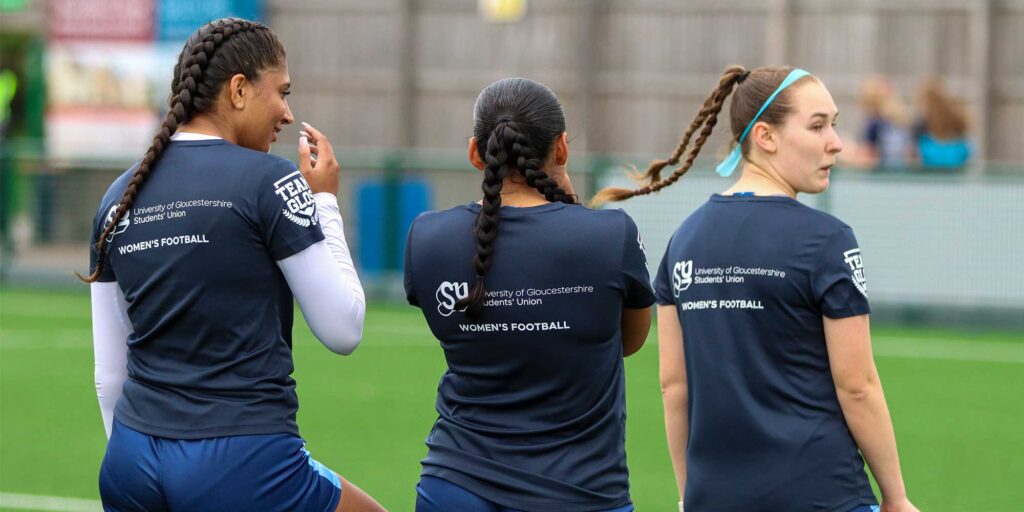 Three female football players with their backs to the camera wearing Team Glos University of Gloucestershire Women's football tshirts showing a Student Union logo.