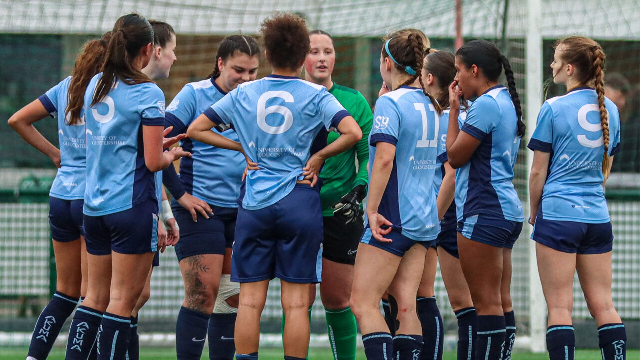 A group of female football players in light blue University of Gloucestershire kits, huddled together on a pitch with a goalpost in the background.