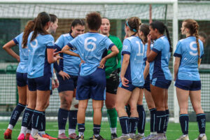 A group of female football players in light blue University of Gloucestershire kits, huddled together on a pitch with a goalpost in the background.