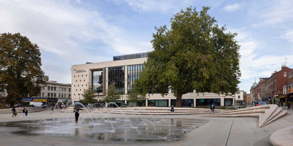 A wide view of the City Campus building, which features large vertical glass windows and “University of Gloucestershire” signage set on the top left side of the building. In the foreground, a ground-level fountain sends water into the air, with several people walking, standing, and one person moving through the water. Large mature trees are featured in the view, and surrounding streets with shops and buildings are visible either side of the building. Above, a blue sky with wispy clouds.
