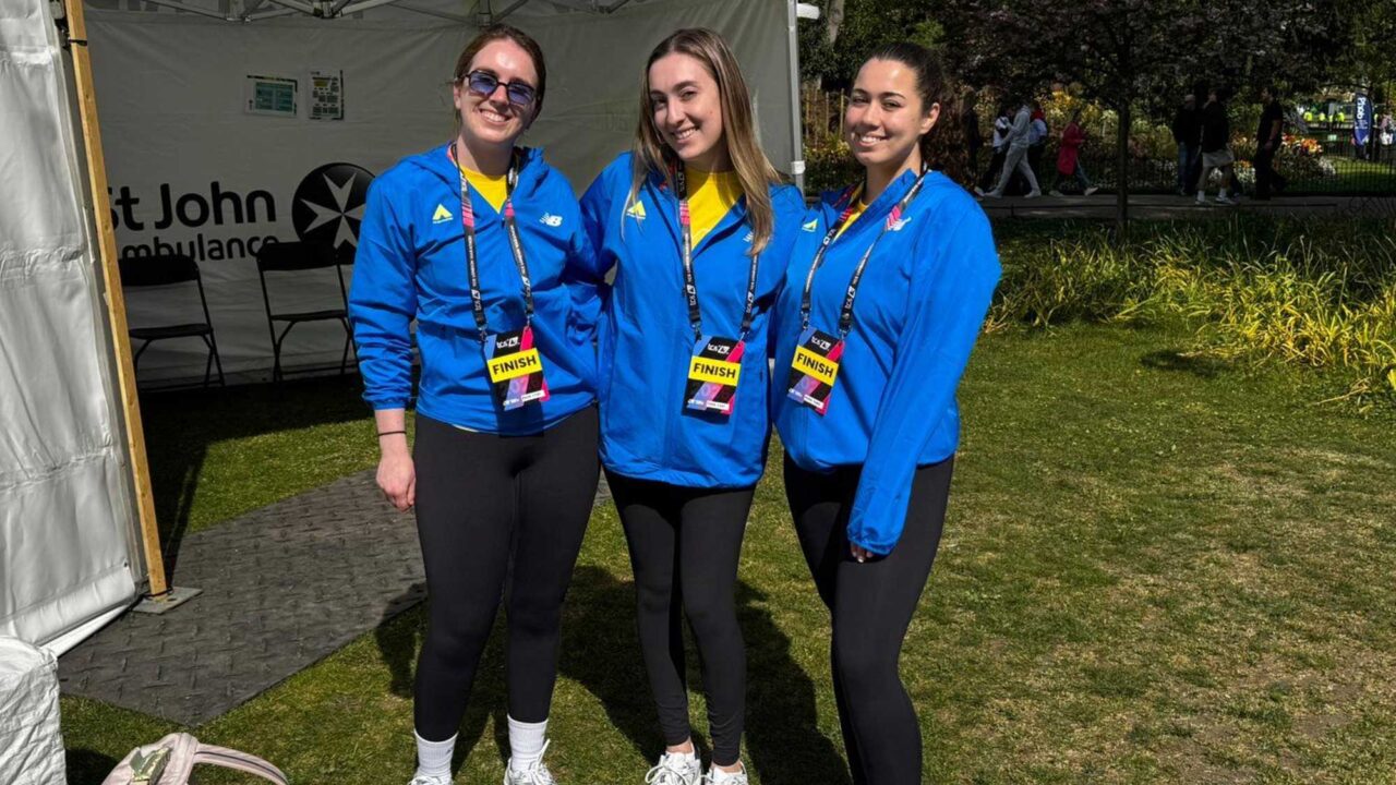 Three Physiotherapy students standing side by side on a grass area, wearing blue jackets, black leggings and trainers, with a St John Ambulance gazebo and trees in the background.