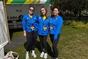 Three Physiotherapy students standing side by side on a grass area, wearing blue jackets, black leggings and trainers, with a St John Ambulance gazebo and trees in the background.