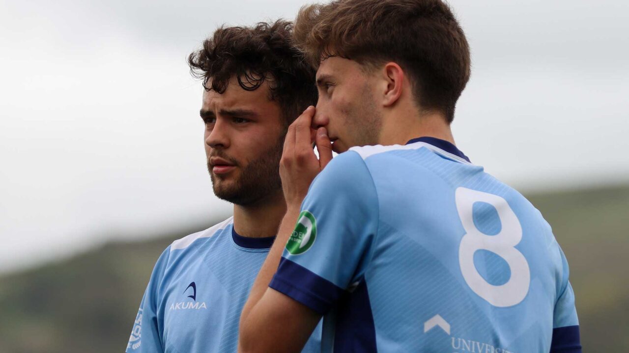Two male football players in University of Gloucestershire football kits, standing close together on the pitch, with one player whispering to the other behind his hand.