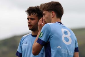 Two male football players in University of Gloucestershire football kits, standing close together on the pitch, with one player whispering to the other behind his hand.
