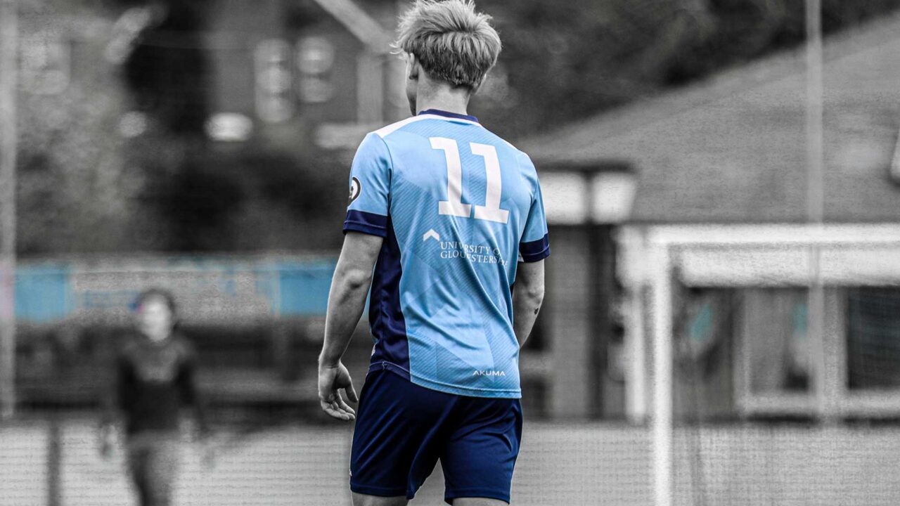 Male football player wearing a light blue University of Gloucestershire kit with the number 11 on the back, standing on a pitch with a blurred goal and another player in the background; the player in colour contrasts against a mostly black-and-white scene.