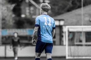 Male football player wearing a light blue University of Gloucestershire kit with the number 11 on the back, standing on a pitch with a blurred goal and another player in the background; the player in colour contrasts against a mostly black-and-white scene.