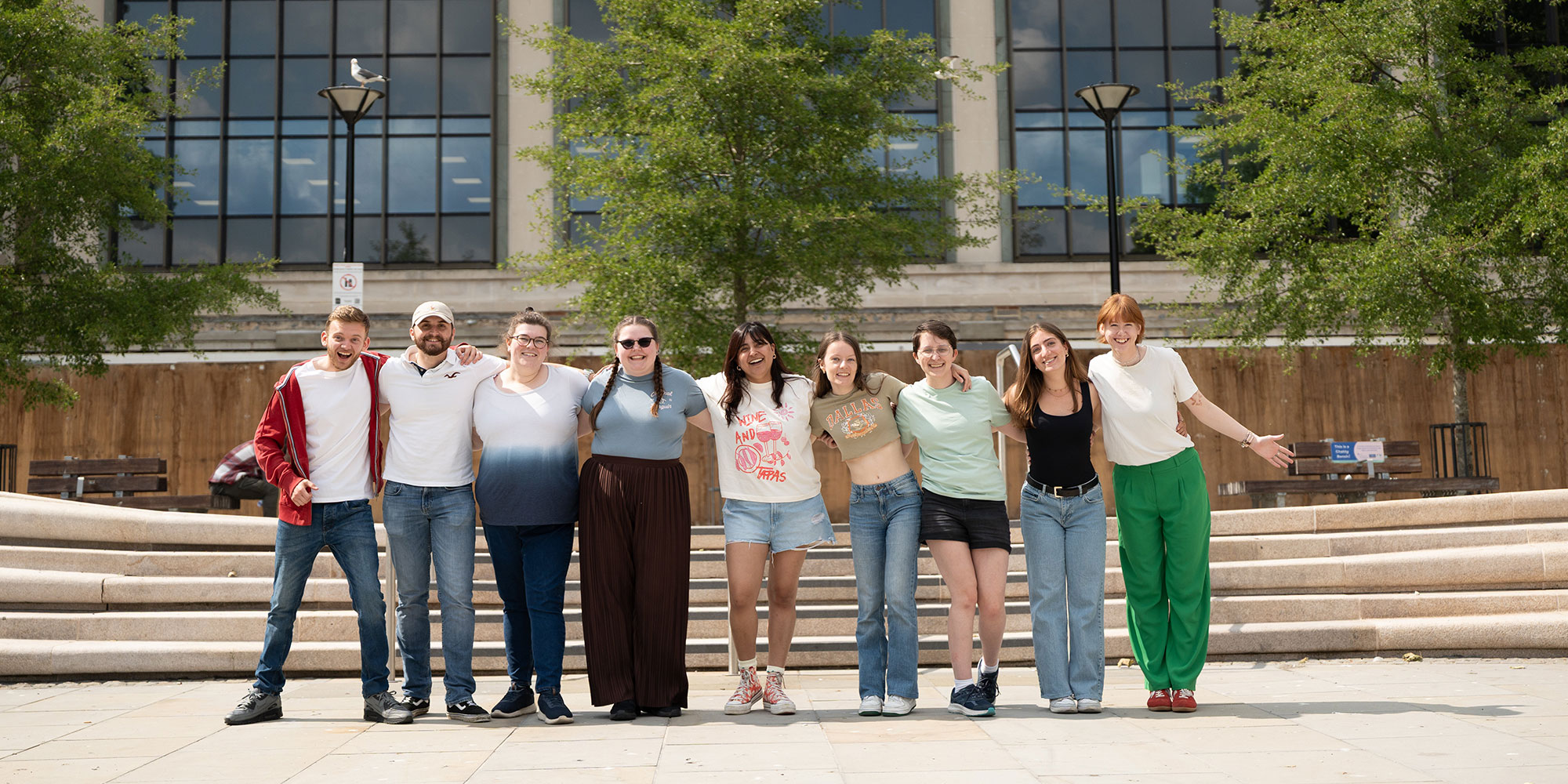 Nine students standing in a row arm in arm and smiling stood in front of steps outside the front of City campus. Trees and benches appear in the background.