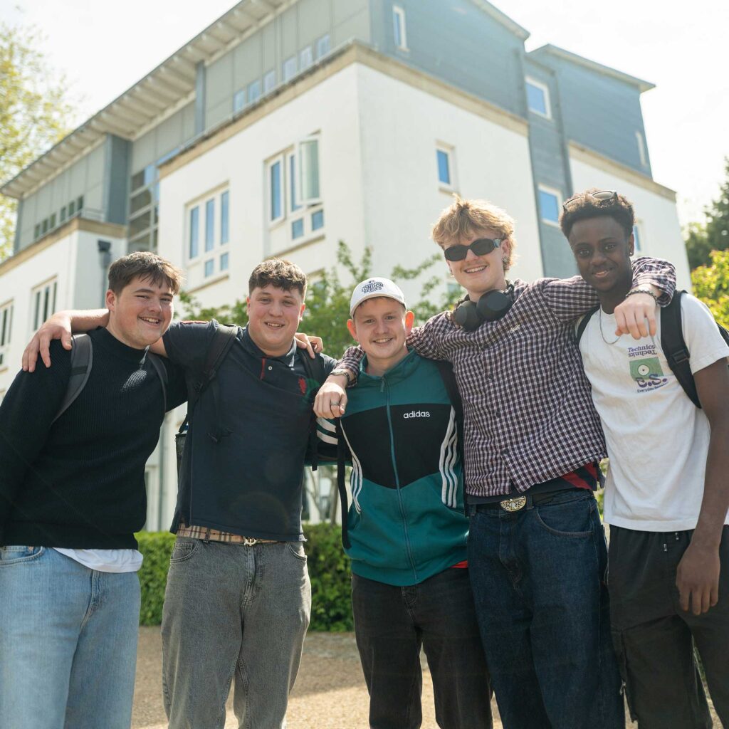 Five male students smiling with arms over each others' shoulders in front of halls accommodation at Park Campus.