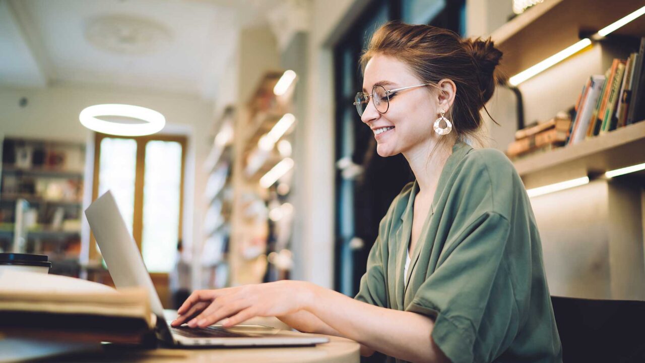 A woman works at a laptop in a workspace with bookshelves.