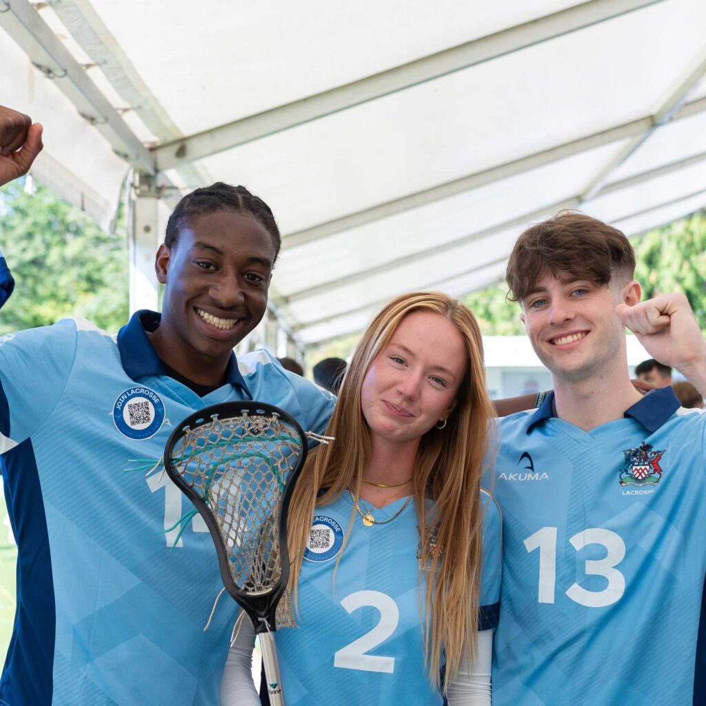 Three members of the lacrosse team wearing light blue University of Gloucestershire kits, the female student in the middle is holding a lacrosse stick.