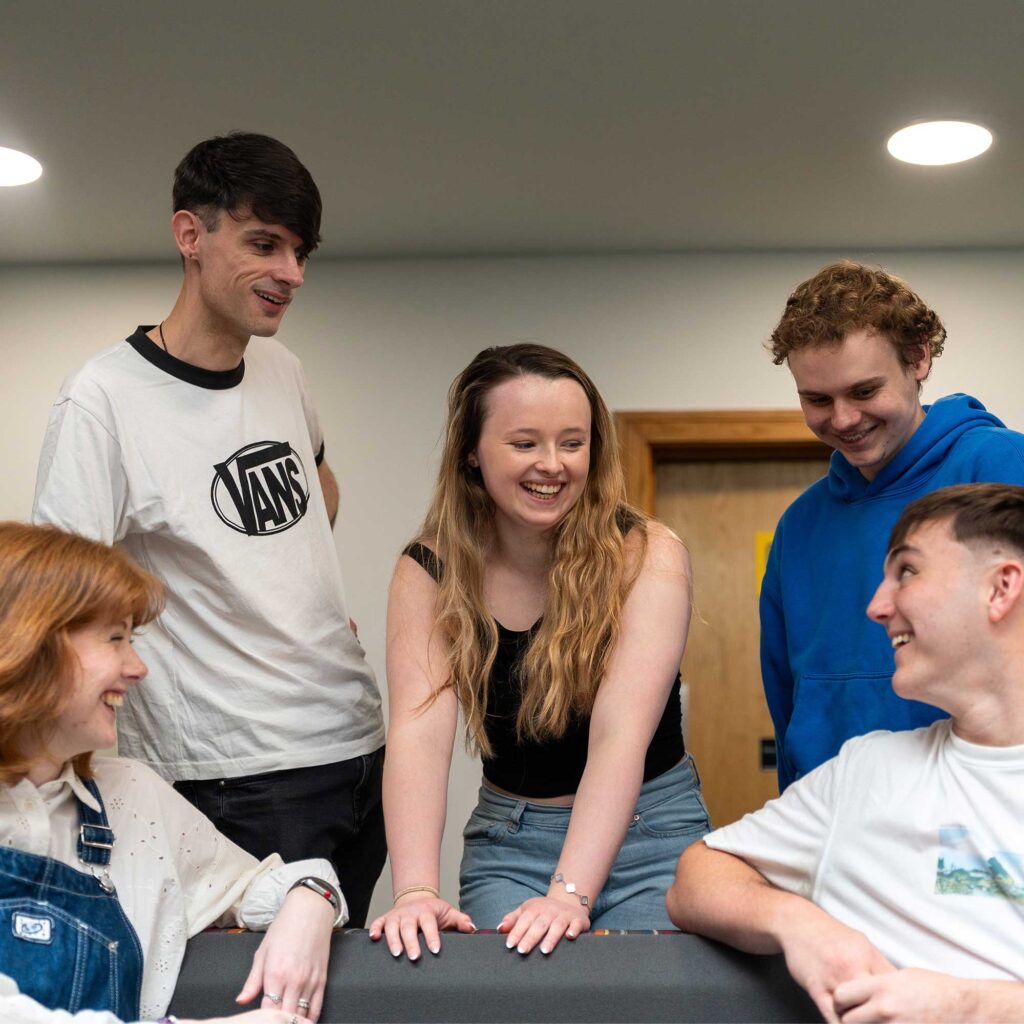 Five students gathered around a seating area indoors, smiling and looking at each other. One person leans forward with hands on the seat. The room has neutral walls, wooden doors, and overhead ceiling lights.