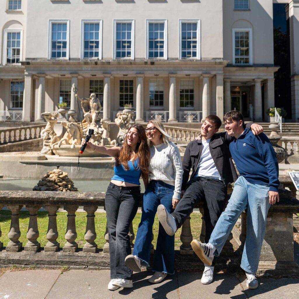 Four students sitting on stone railings on a sunny day, posing for a selfie, in front of a large building with tall windows and columns, and a decorative fountain.