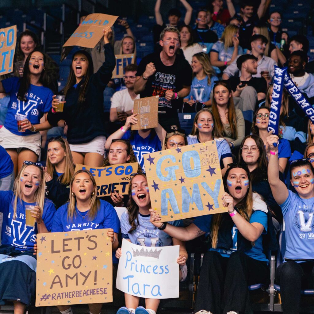 A indoor arena of spectators cheering and holding handmade signs such as “GO AMY,” “LET’S GO AMY,” and “Princess Tara.” Several people raise foam sticks and drinks.