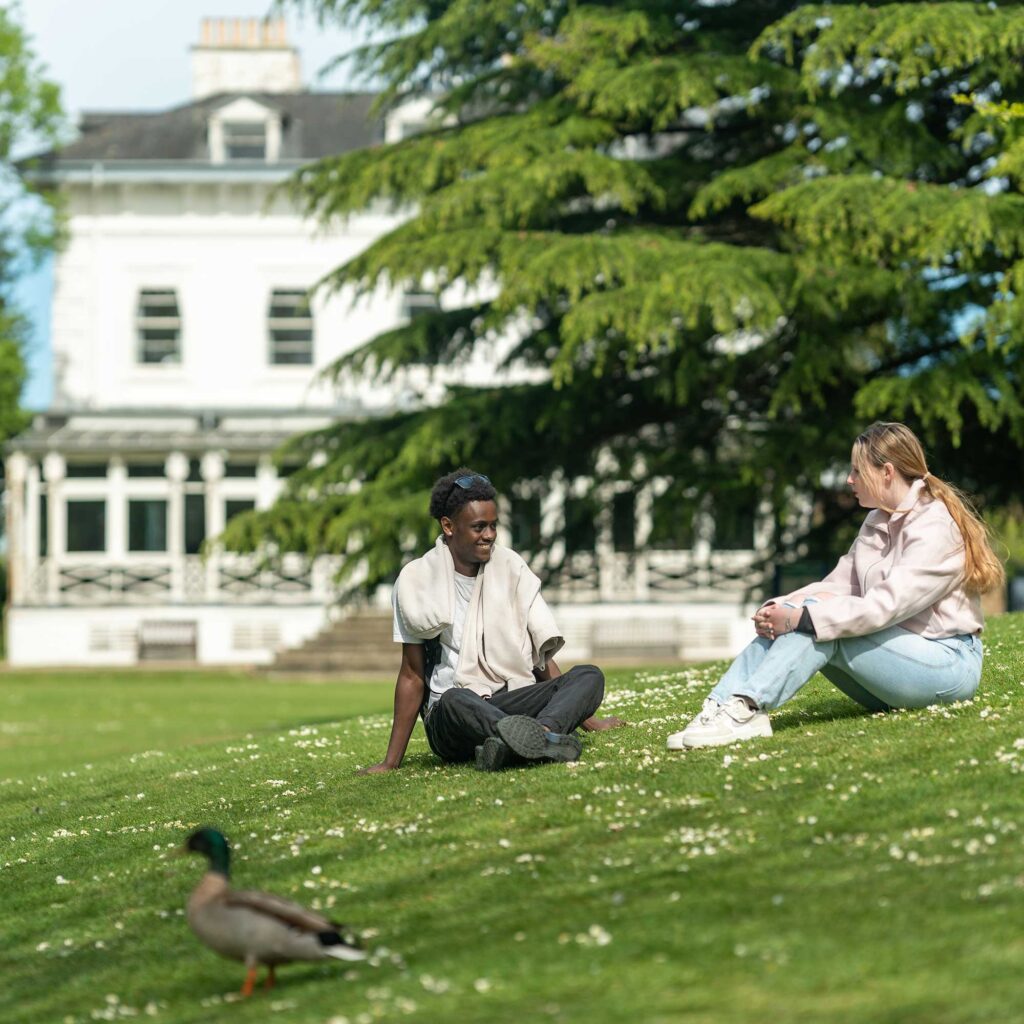 Two students sitting on the grass at Park Campus with a university building behind them.