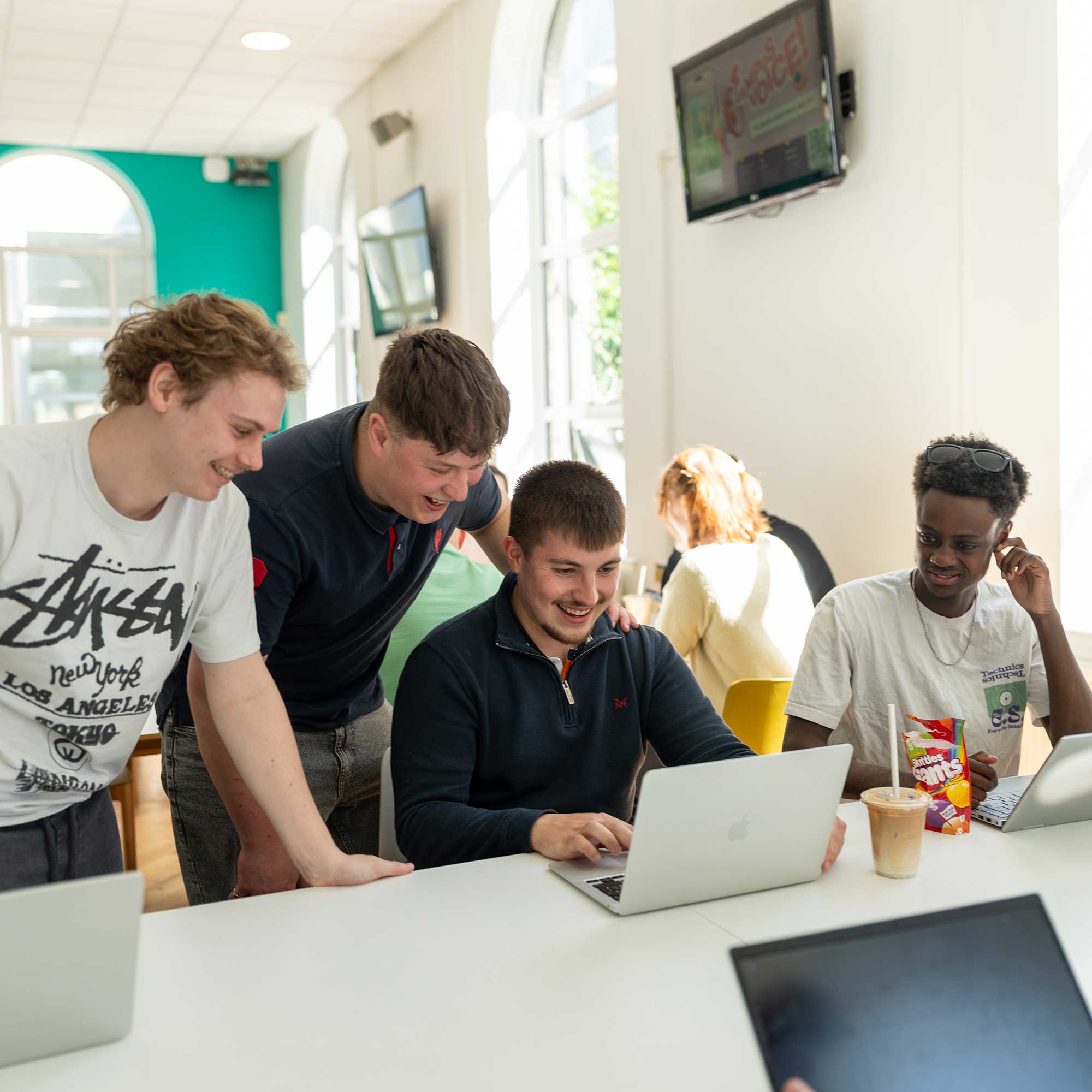 A group of students in the refectory look at a laptop screen toegther.