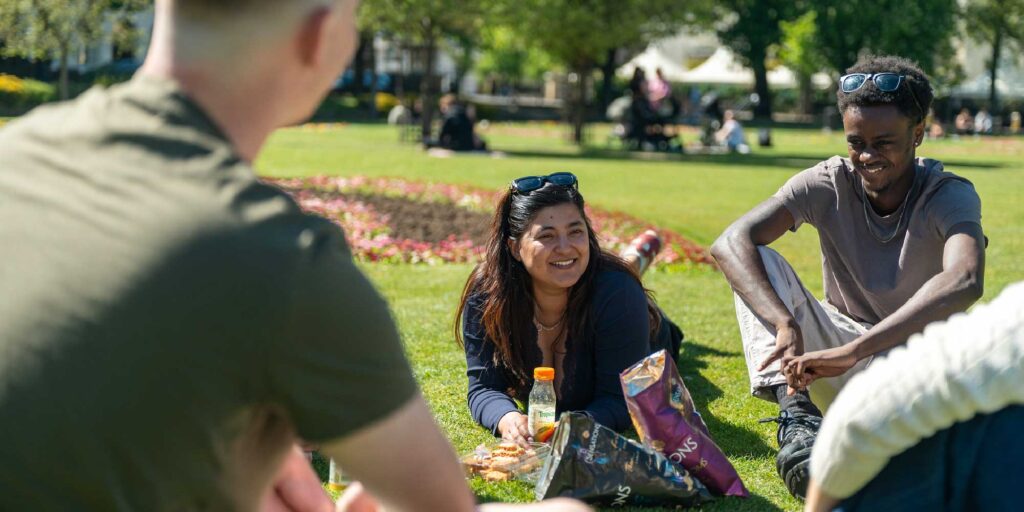 Students lying on the grass in a park on a sunny day with food and drinks.