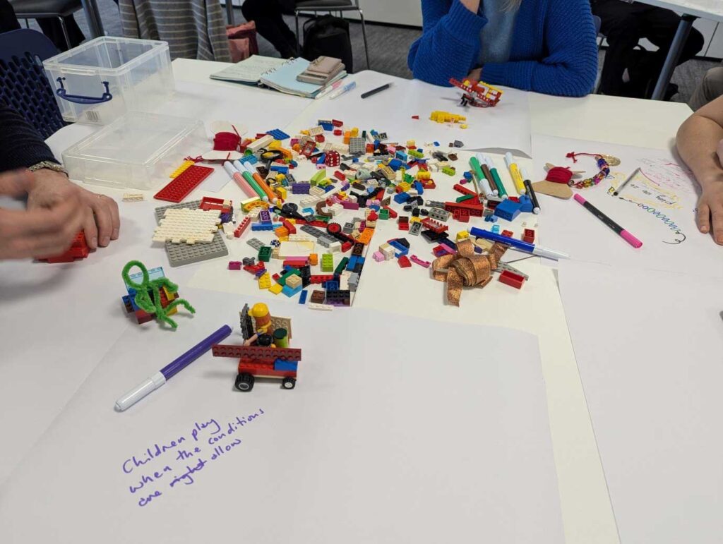 A close up of various items on a table including Lego, pens and other items, with people sat at the table.